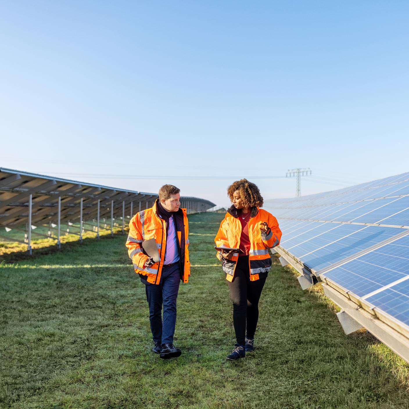 Zwei Techniker in reflektierender Kleidung gehen zwischen den Reihen von Photovoltaikmodulen in einem Solarpark.