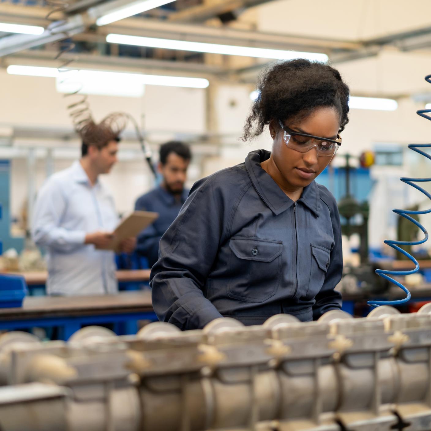 Mulher jovem afro-americana trabalhando em uma produção de montagem de bombas de água em uma fábrica - Pessoas incidentais ao fundo