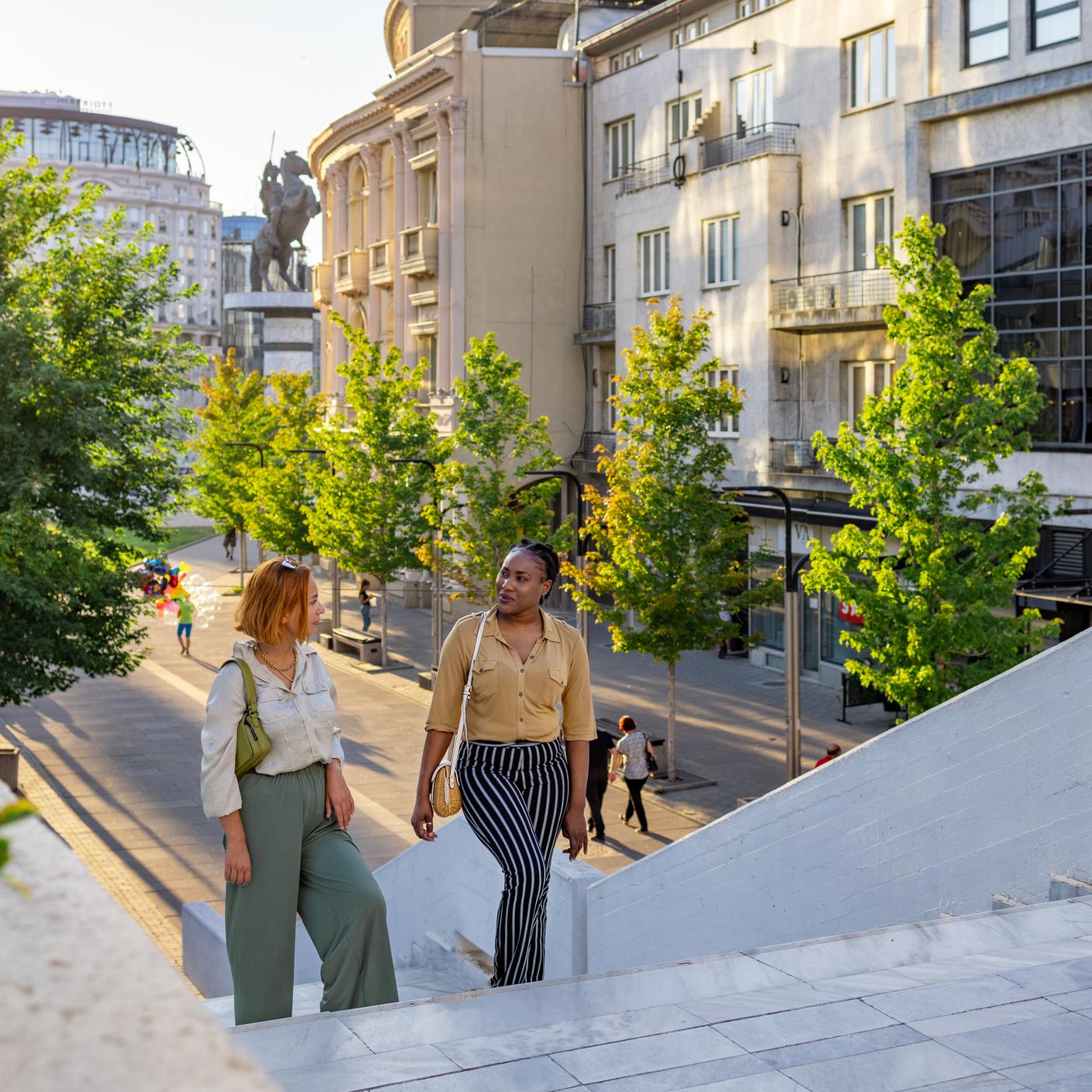 Dos amigas paseando por una calle urbana, rodeadas de gente, edificios modernos y vegetación.