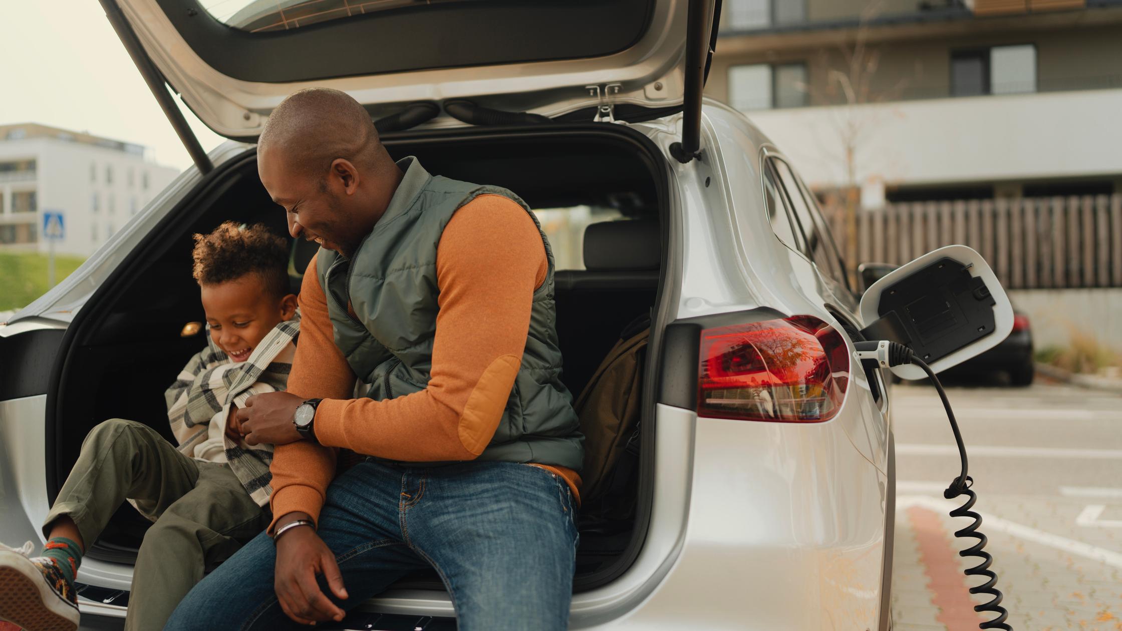 Sustainability in Transport and Mobility -  Multiracial father with his son sitting in car trunk and waiting while their electric car charging