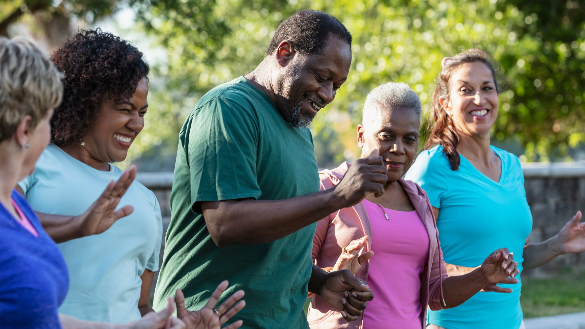 A multiracial group of five adults in a dance exercise class at the park, standing in a row, side by side.