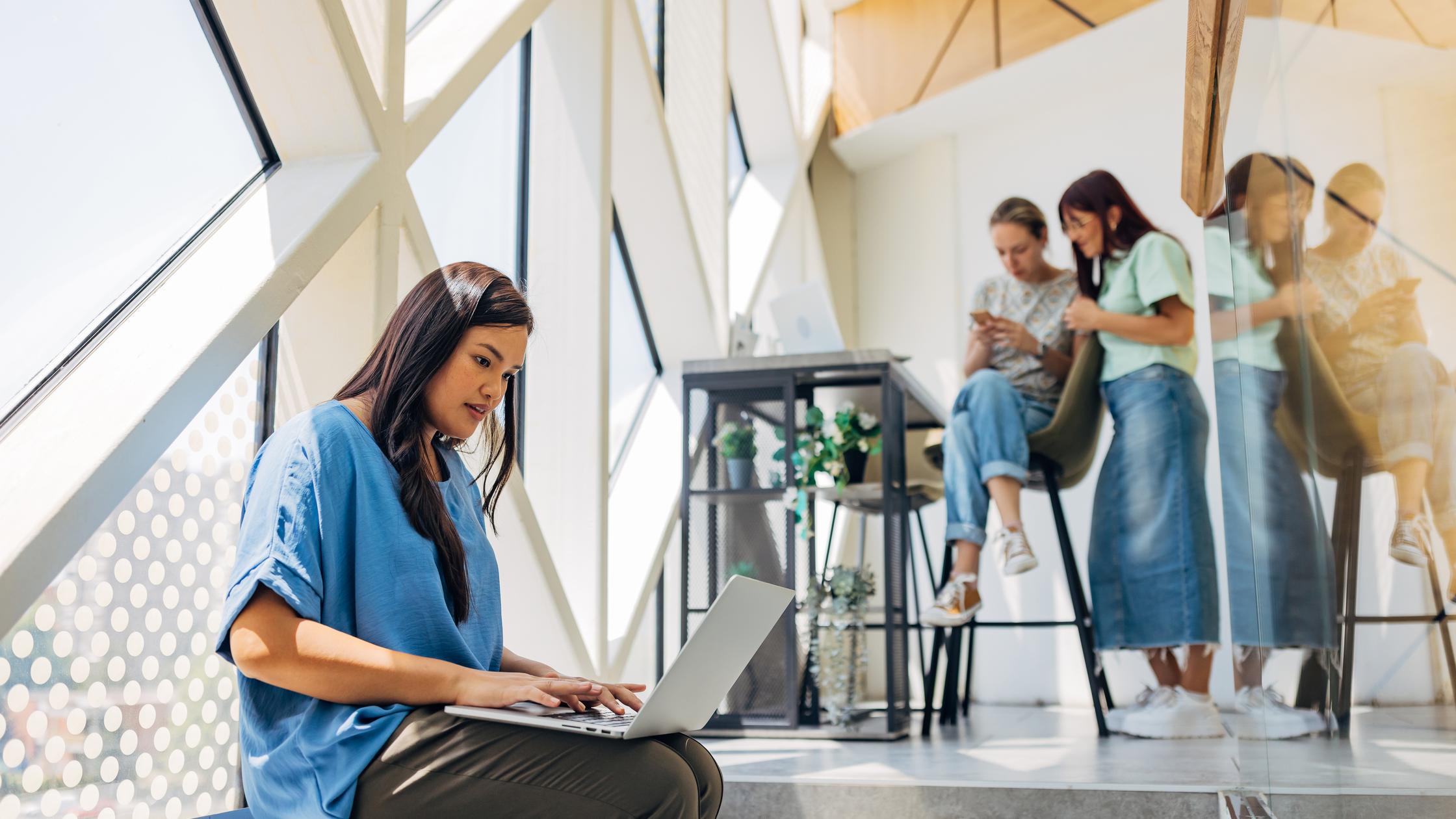 Femme d'affaires sur son ordinateur portable, assise non loin de ses collègues dans un environnement professionnel hybride